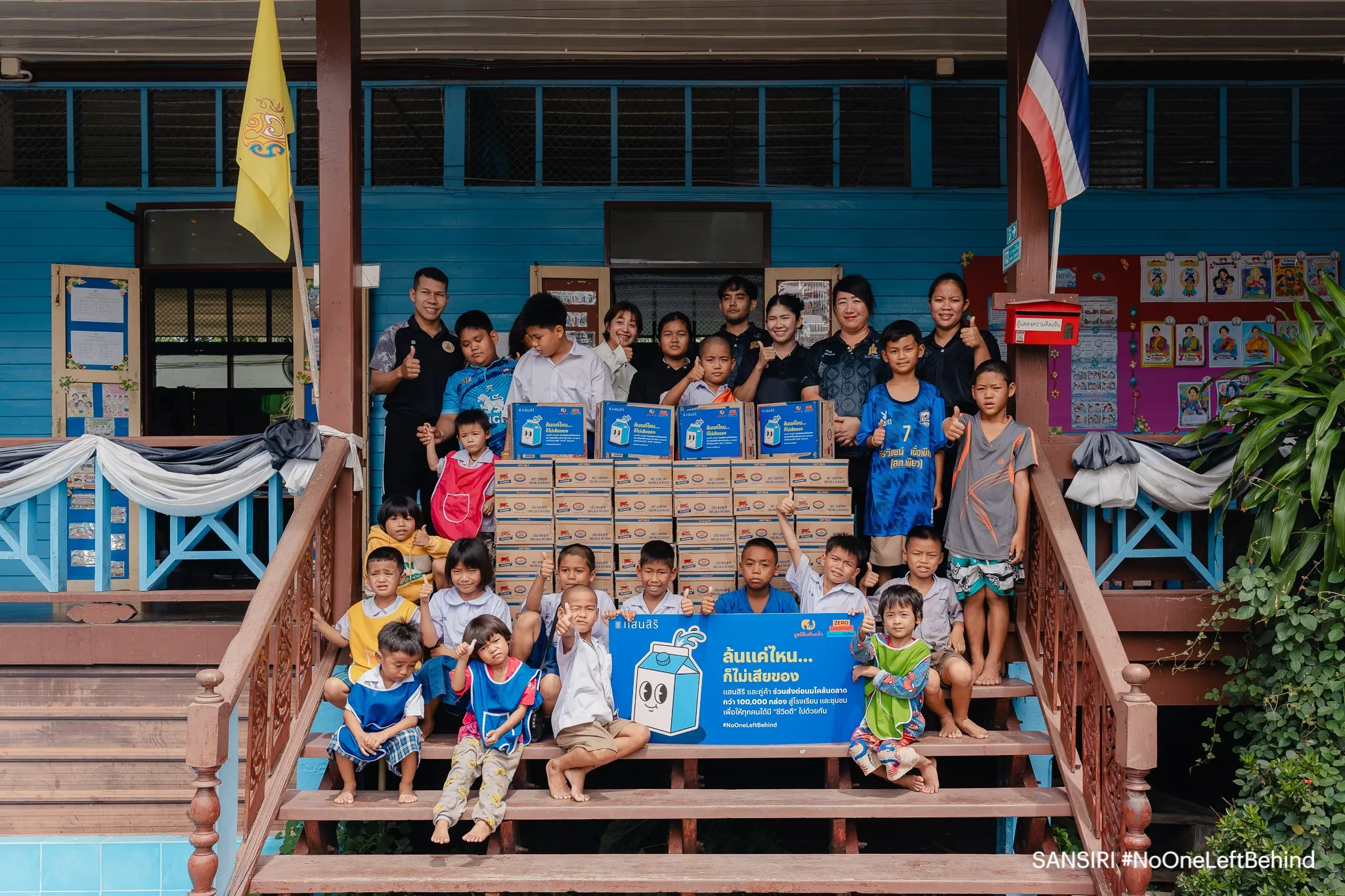 กลุ่มเด็กนักเรียนและเจ้าหน้าที่ถ่ายภาพร่วมกันบนบันไดโรงเรียน พร้อมกล่องนมจำนวนมากและป้ายกิจกรรม #NoOneLeftBehind ของแสนสิริที่ส่งมอบนมให้แก่โรงเรียนในโครงการช่วยเหลือเกษตรกรโคนม
