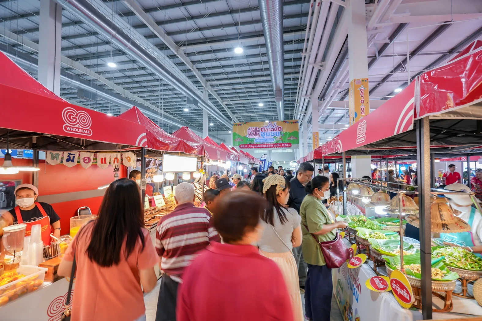 ภาพบรรยากาศตลาดนัดชุมชนภายในโก โฮลเซลล์ ที่มีผู้คนจำนวนมากเดินเลือกซื้อสินค้าจากร้านค้าต่างๆ ใต้ผ้าใบสีแดงของบูธ SME