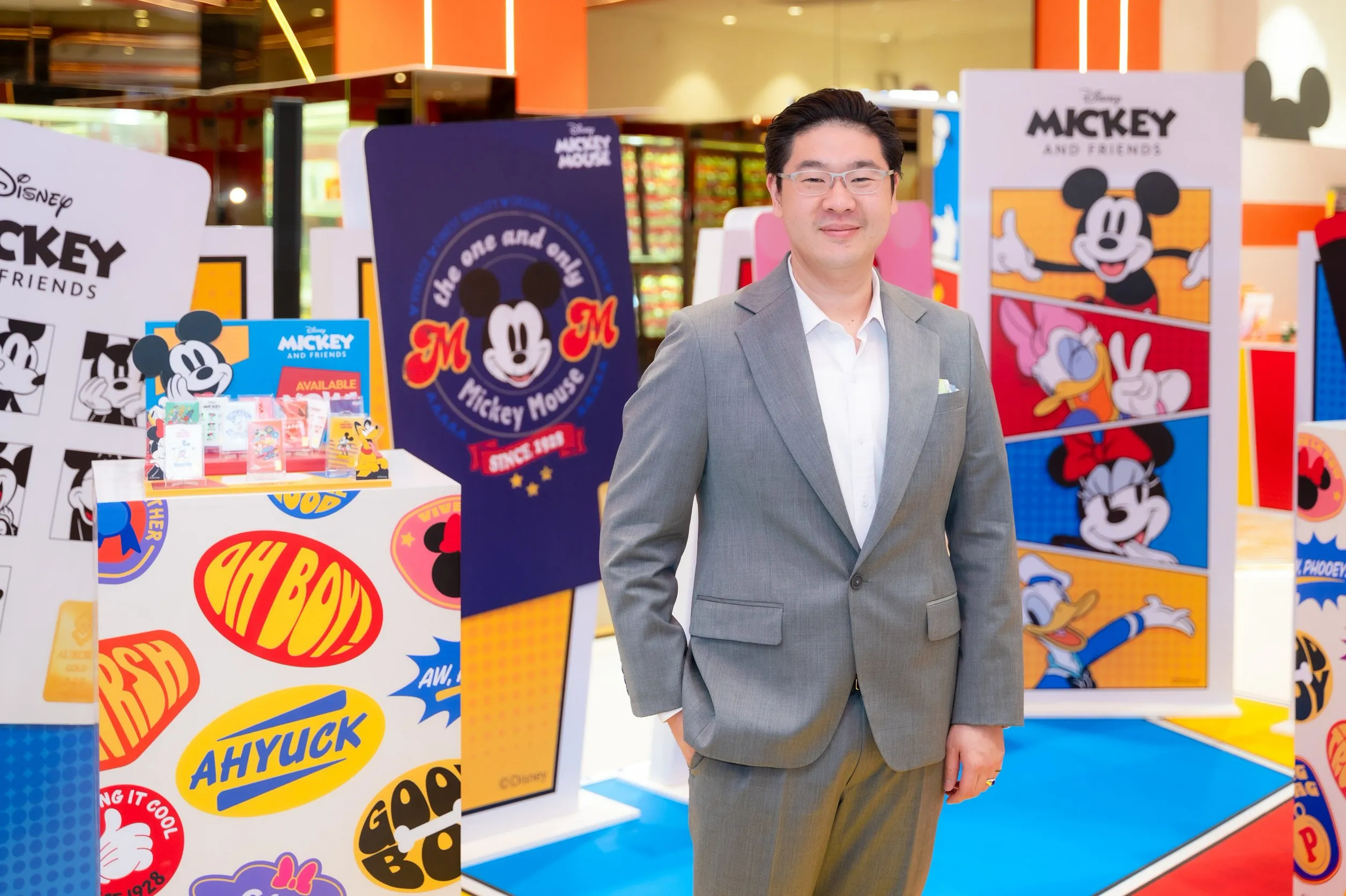 ผู้บริหารสวมสูทสีเทายืนยิ้มถ่ายรูปท่ามกลางบูธจัดแสดงสินค้าคอลเลกชัน Disney Mickey and Friends ที่มีลวดลายมิกกี้เมาส์และตัวการ์ตูนดิสนีย์หลากสีสัน