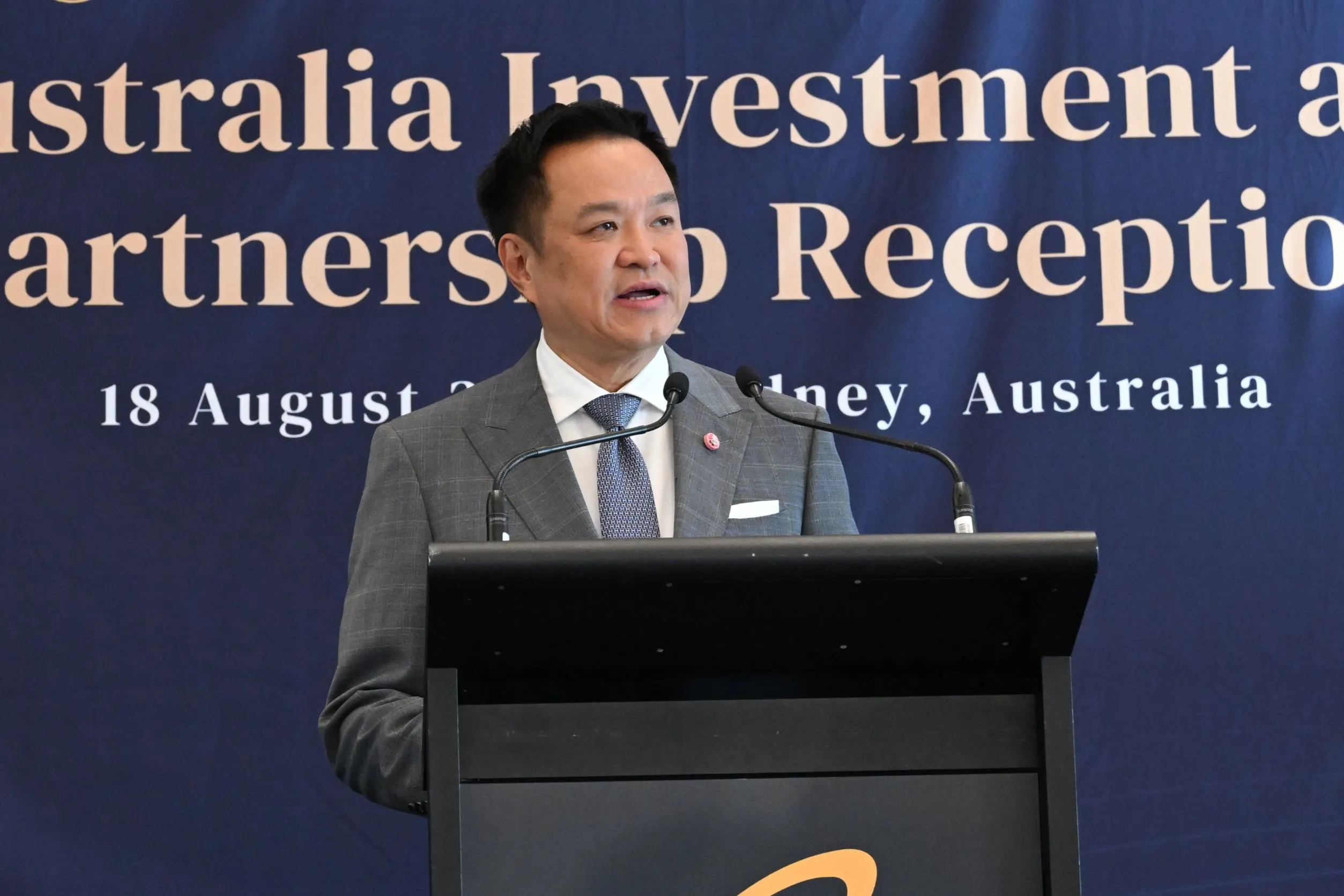 ชายสวมสูทสีเทากำลังกล่าวสุนทรพจน์ที่แท่นพูดในงาน Australia Investment and Partnership Reception ซึ่งจัดขึ้นเมื่อวันที่ 18 สิงหาคม ณ ประเทศออสเตรเลีย โดยมีป้ายพื้นหลังสีน้ำเงินเข้มเป็นฉากหลัง