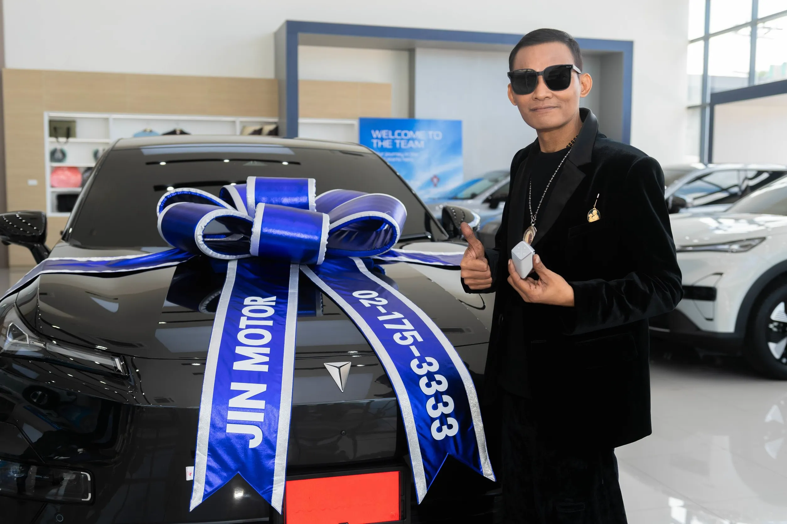ชายสวมสูทและแว่นตาดำแสดงท่าชูนิ้วโป้งข้างรถยนต์ไฟฟ้าสีดำที่ประดับโบว์สีน้ำเงินของจิน มอเตอร์ภายในโชว์รูมรถยนต์