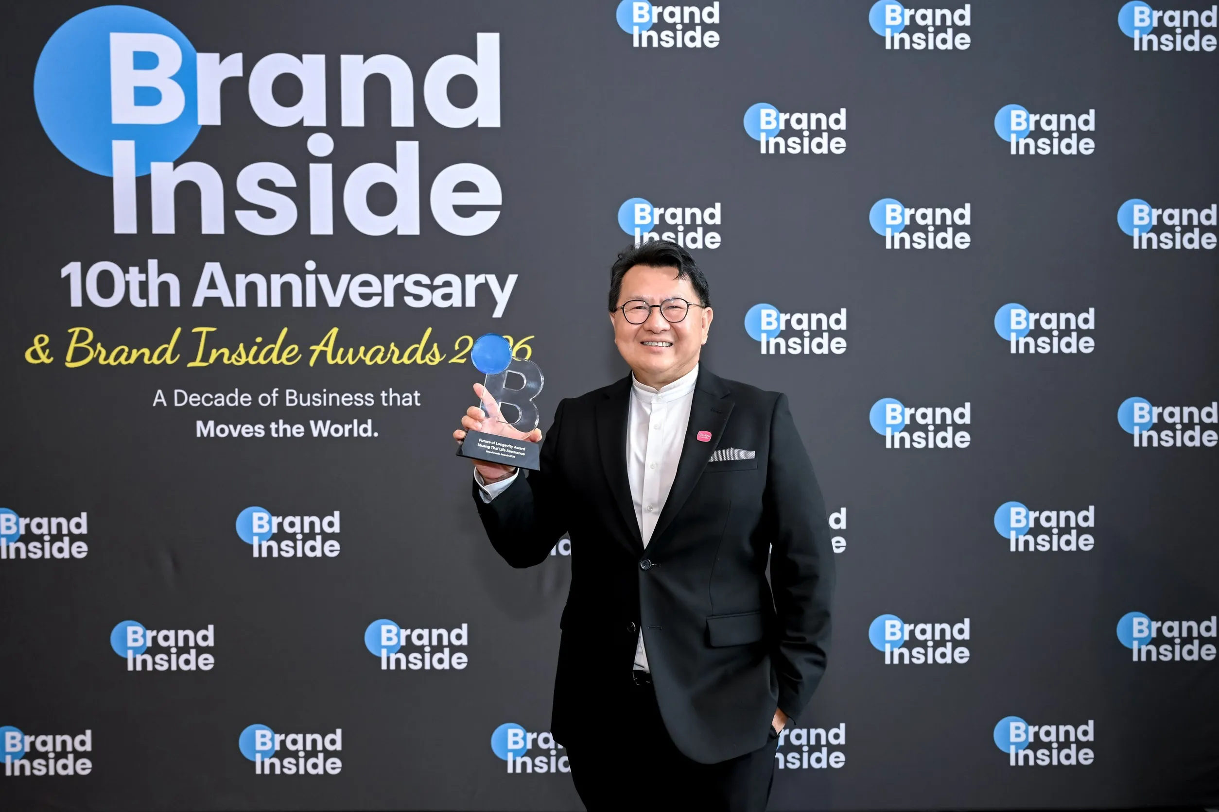 ผู้บริหารเมืองไทยประกันชีวิตสวมสูทสีดำถือถ้วยรางวัล Future of Longevity Award ยืนยิ้มถ่ายภาพหน้าแบ็คดรอปงาน Brand Inside 10th Anniversary & Brand Inside Awards 2026