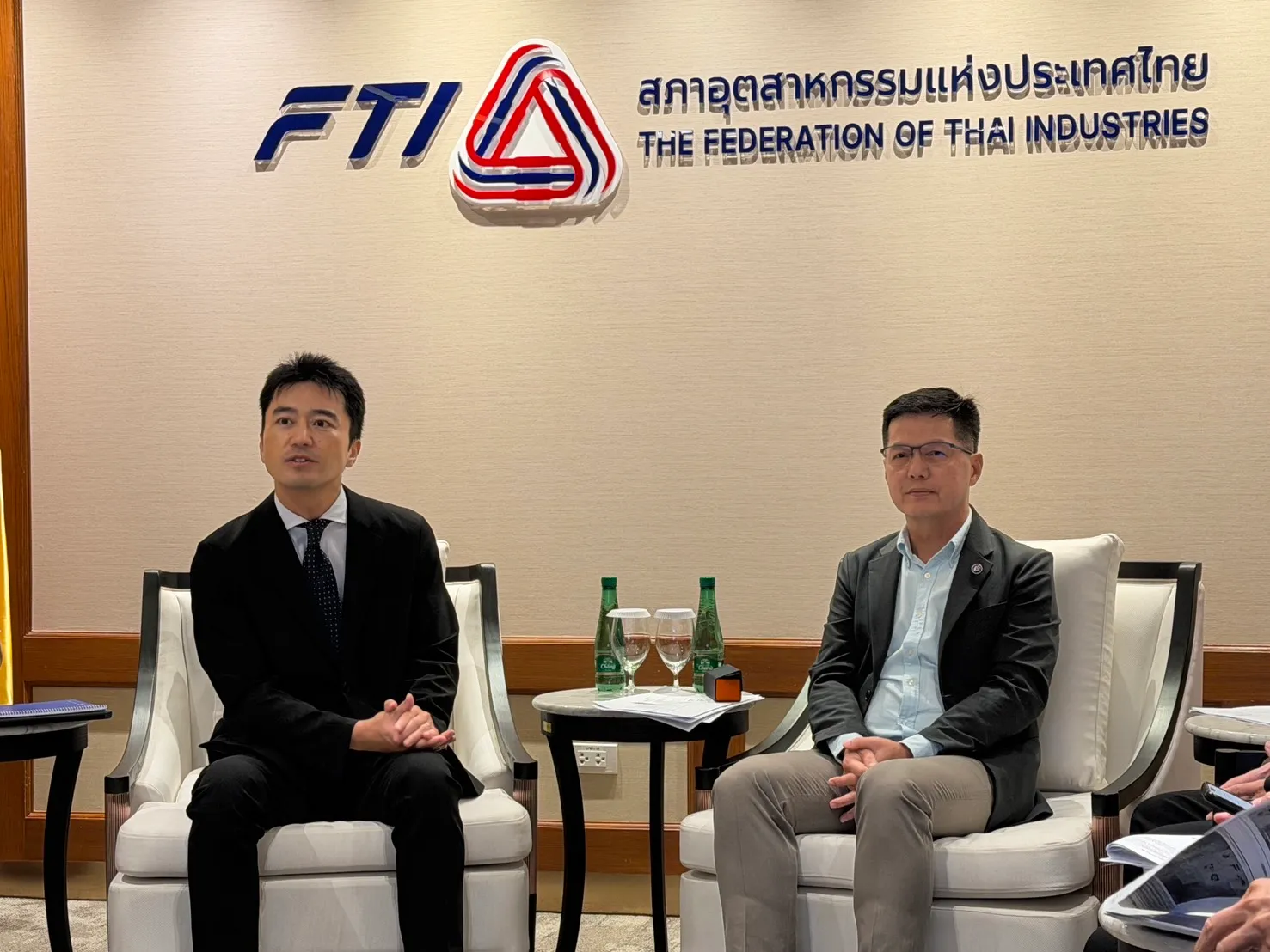 ผู้บริหารสองท่านนั่งสนทนาบนเก้าอี้สีขาวในห้องประชุมของสภาอุตสาหกรรมแห่งประเทศไทย โดยมีโต๊ะวางน้ำดื่มอยู่ตรงกลาง และมีโลโก้ FTI ติดอยู่บนผนังด้านหลัง