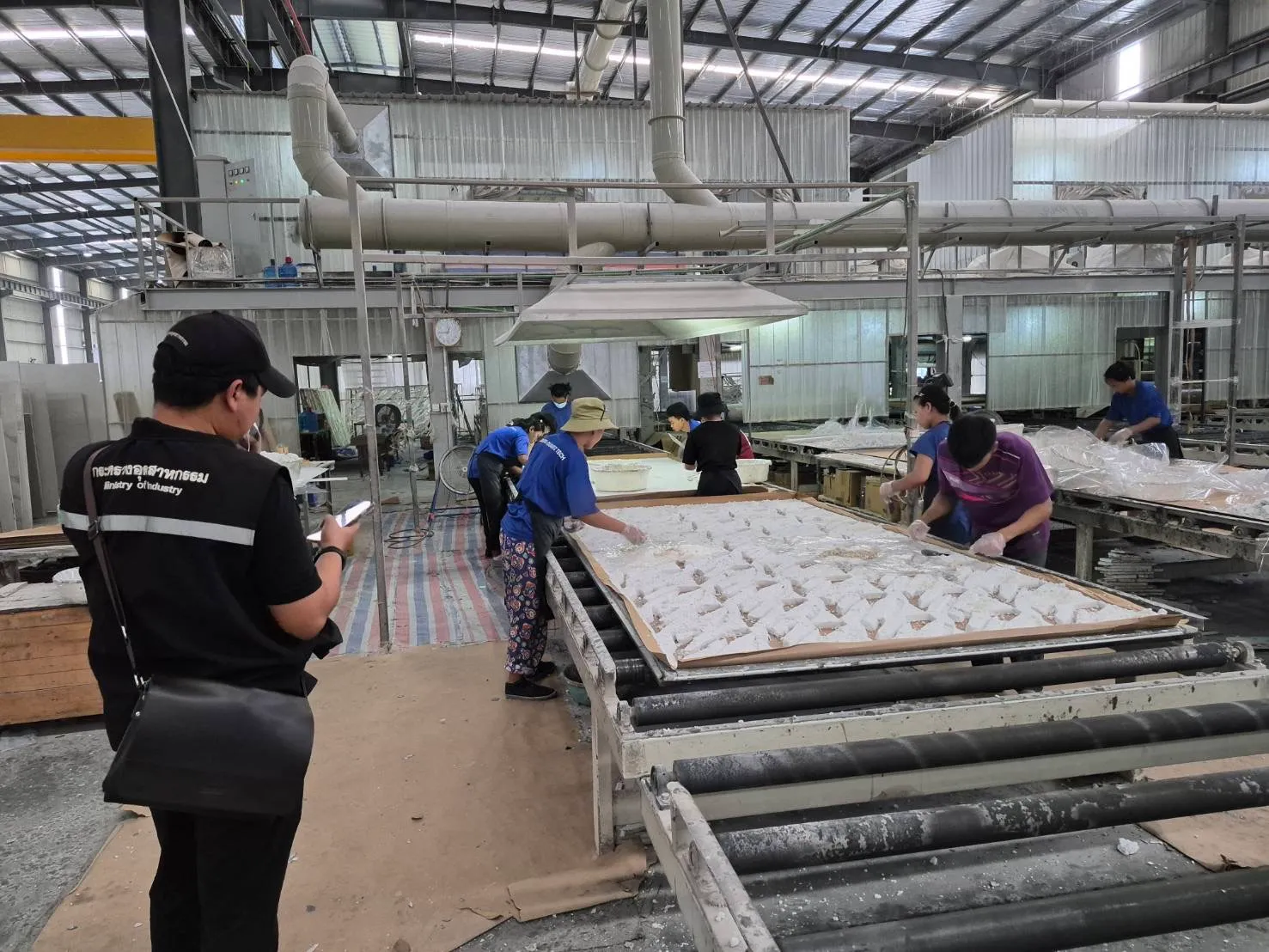 เจ้าหน้าที่กระทรวงอุตสาหกรรมถ่ายภาพขณะลงพื้นที่ตรวจสอบกระบวนการผลิตของโรงงานพนัสนิคม โดยมีคนงานกำลังปฏิบัติงานบนสายพานลำเลียงวัสดุในโรงงาน