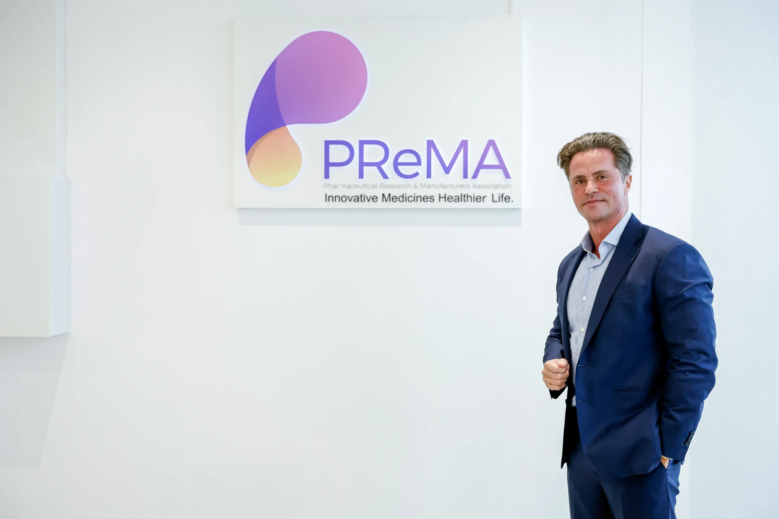 ผู้ชายสวมสูทสีกรมท่ายืนยิ้มถ่ายภาพข้างป้ายโลโก้ PReMA (Pharmaceutical Research & Manufacturers Association) ที่มีข้อความ "Innovative Medicines Healthier Life" ติดอยู่บนผนังสีขาว