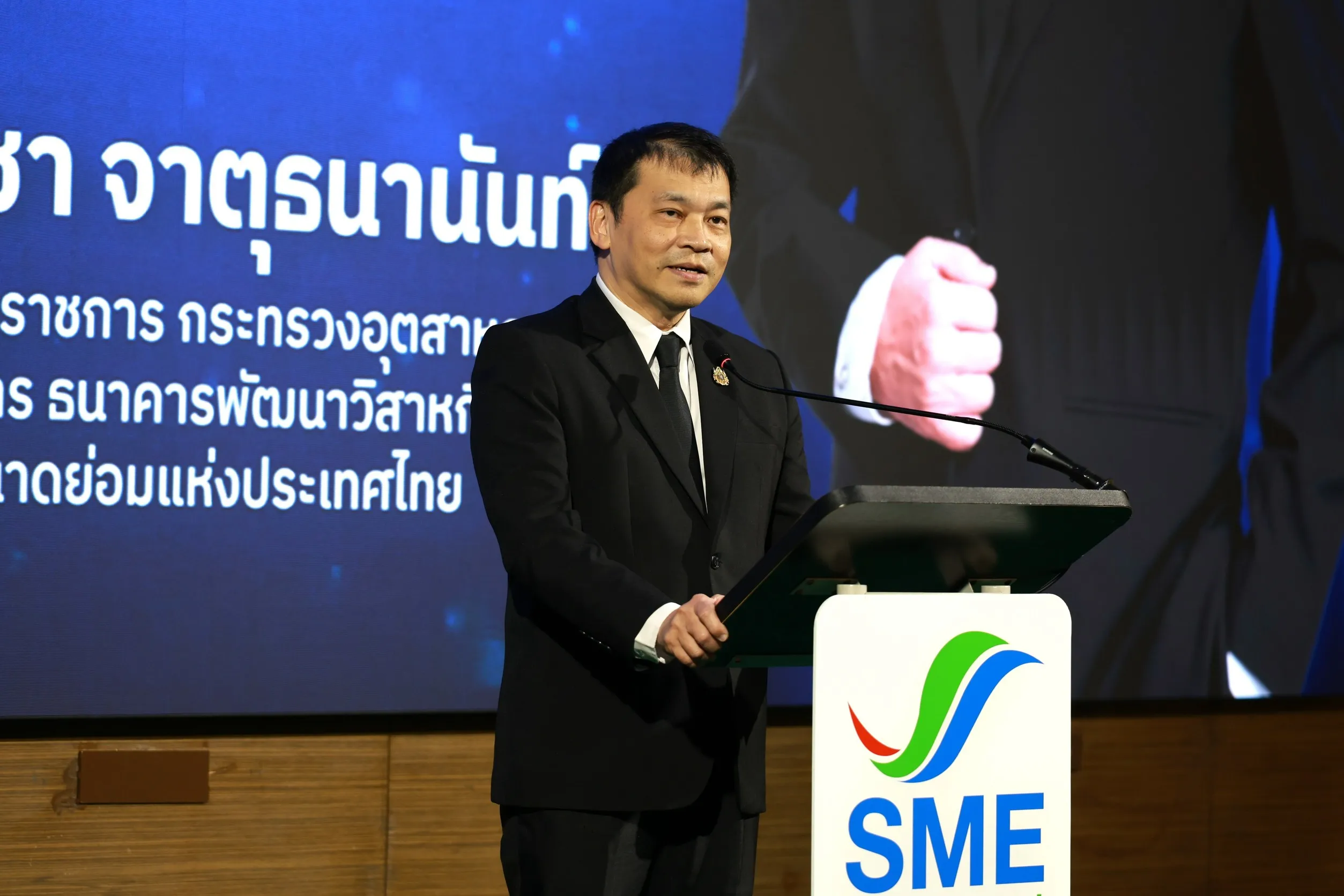 ผู้บริหารสวมสูทสีดำกำลังกล่าวสุนทรพจน์บนเวทีที่แท่นพูดโลโก้ SME หน้าจอพื้นหลังสีน้ำเงินแสดงข้อความภาษาไทยเกี่ยวกับกระทรวงอุตสาหกรรมและธนาคารพัฒนาวิสาหกิจขนาดกลางและขนาดย่อมแห่งประเทศไทย
