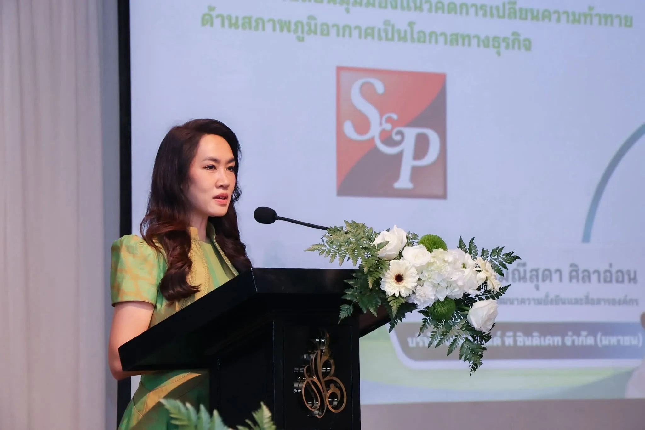 ผู้บริหารหญิงในชุดไทยสีเขียวกำลังกล่าวปราศรัยที่หน้าไมโครโฟนบนโพเดียม โดยมีดอกไม้สีขาวประดับและโลโก้ S&P ปรากฏอยู่บนแบ็คดรอปด้านหลัง ในงานแถลงข่าวการรับรอง Carbon Neutral Product
