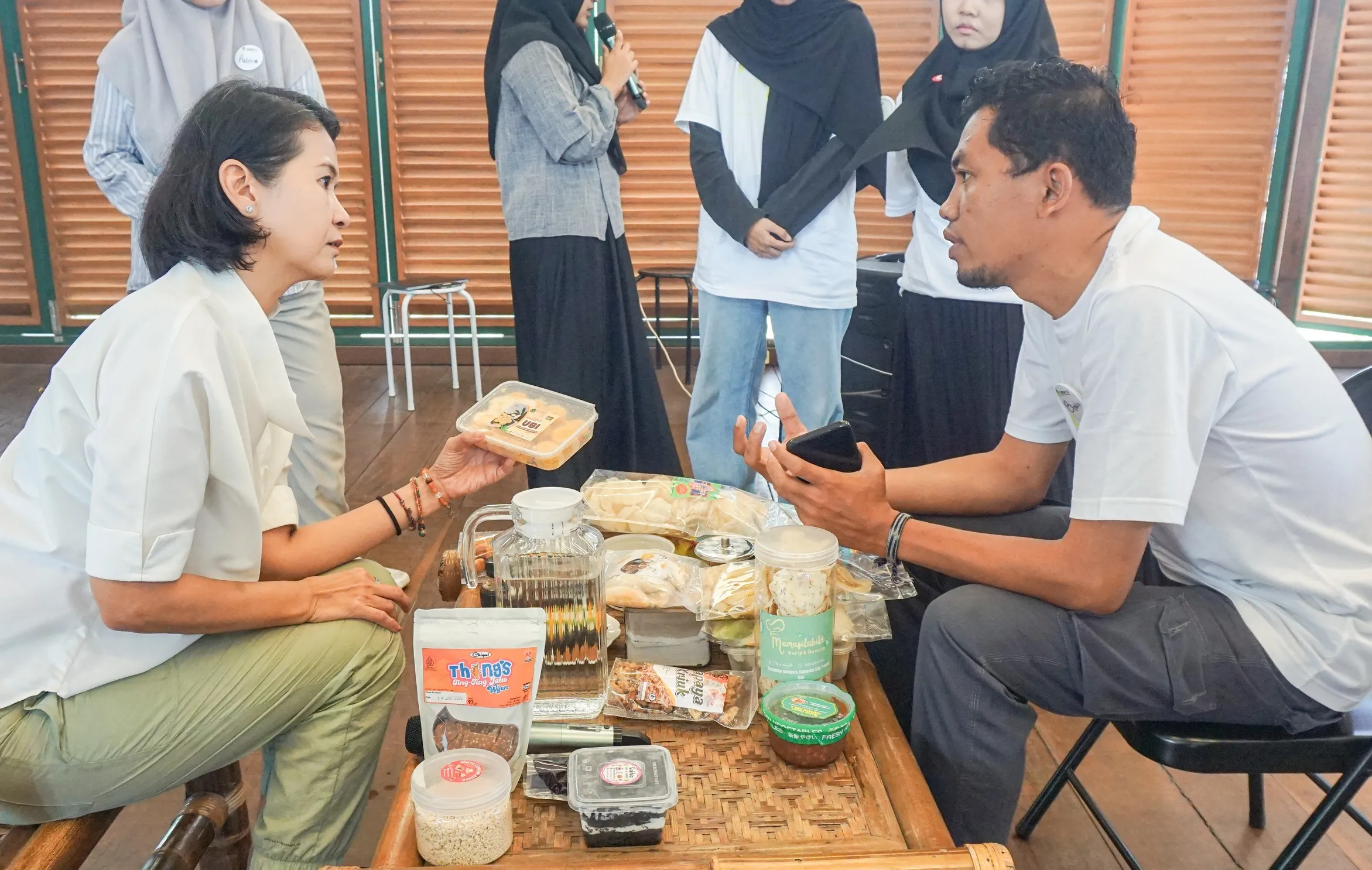 ภาพผู้หญิงกำลังพูดคุยและรับผลิตภัณฑ์อาหารแปรรูปจากชายคนหนึ่งที่นั่งอยู่ด้านหลังโต๊ะซึ่งเต็มไปด้วยผลิตภัณฑ์อาหารหลากหลายชนิด โดยมีกลุ่มคนยืนอยู่ด้านหลังในบรรยากาศงานแสดงสินค้าหรือกิจกรรมชุมชน