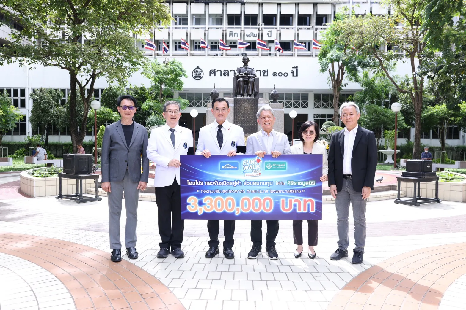 ภาพผู้บริหารโฮมโปรและคณะผู้บริหารโรงพยาบาลศิริราช 6 ท่าน ถ่ายรูปร่วมกันพร้อมป้ายส่งมอบเงินบริจาค 3,300,000 บาท สมทบกองทุนปรับปรุงห้องผ่าตัดตึกสยามินทร์ โรงพยาบาลศิริราช จากโครงการ HomePro Fun Walk 2026 บริเวณลานหน้าตึกศาลาศิริราช 100 ปี