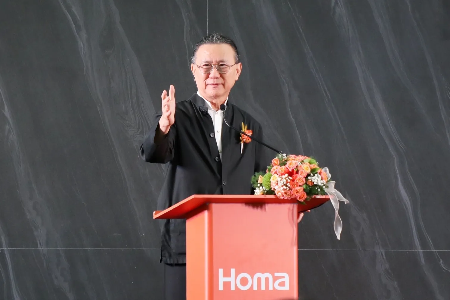 ชายสูงอายุสวมชุดสีดำกำลังกล่าวสุนทรพจน์บนเวทีที่แท่นพูดสีส้มแบรนด์ Homa ซึ่งมีดอกไม้ประดับตกแต่ง ในงานเปิดตัวโครงการลงทุนผลิตตู้เย็นของบริษัทจีนในนิคมอุตสาหกรรมอมตะ
