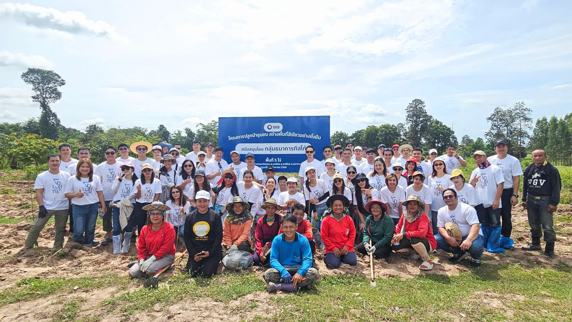 กลุ่มพนักงานจิตอาสาทิสโก้สวมเสื้อ Volunteer สีขาวถ่ายภาพร่วมกับชาวบ้านในชุมชนที่หน้าป้ายโครงการปลูกป่าชุมชน จ.มหาสารคาม ท่ามกลางพื้นที่สีเขียวและต้นไม้กลางแจ้ง
