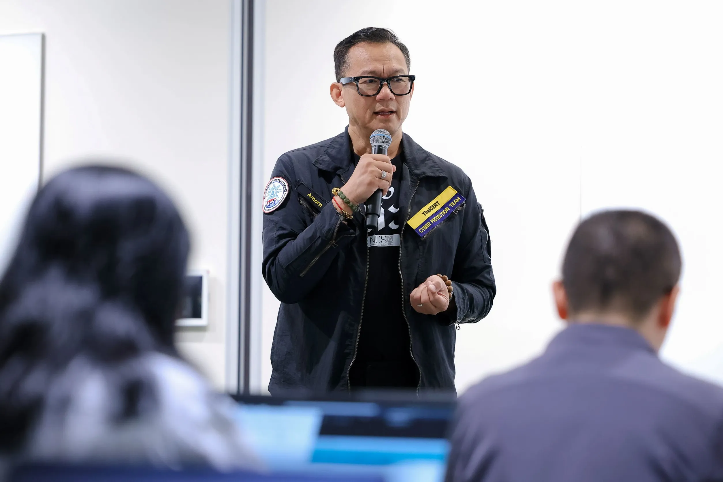 ชายสวมแว่นตาและเสื้อแจ็คเก็ตสีเข้มติดป้าย ThaiCERT Cyber Protection Team กำลังถือไมโครโฟนบรรยายให้ผู้ฟังในงานสัมมนาความมั่นคงปลอดภัยไซเบอร์