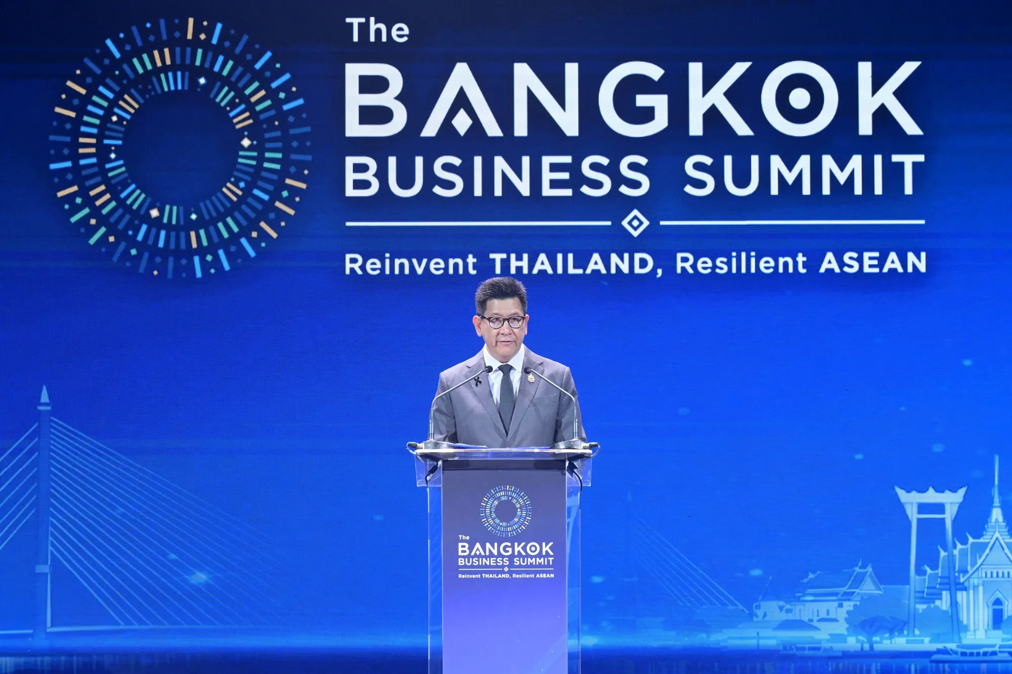 ชายสวมสูทสีเทายืนกล่าวสุนทรพจน์หลังแท่นพูดบนเวทีงาน The Bangkok Business Summit ภายใต้แนวคิด Reinvent Thailand, Resilient ASEAN พื้นหลังสีน้ำเงินมีโลโก้งานและภาพเงาสถาปัตยกรรมกรุงเทพมหานคร