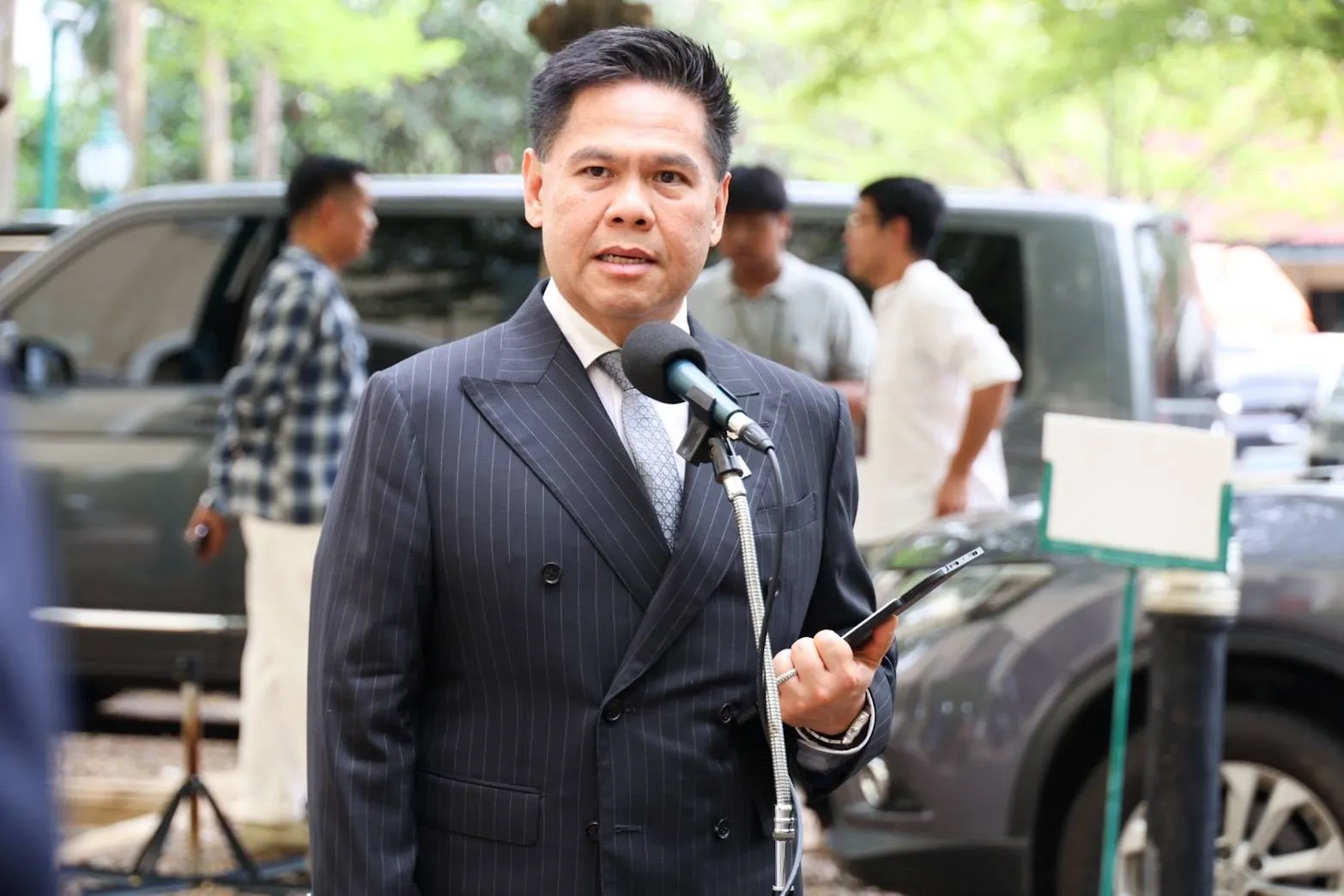 ชายสวมสูทลายทางสีเข้มกำลังพูดผ่านไมโครโฟนขณะถือโทรศัพท์มือถือในมือ โดยมีรถยนต์และผู้คนอยู่ด้านหลังในบรรยากาศกลางแจ้ง