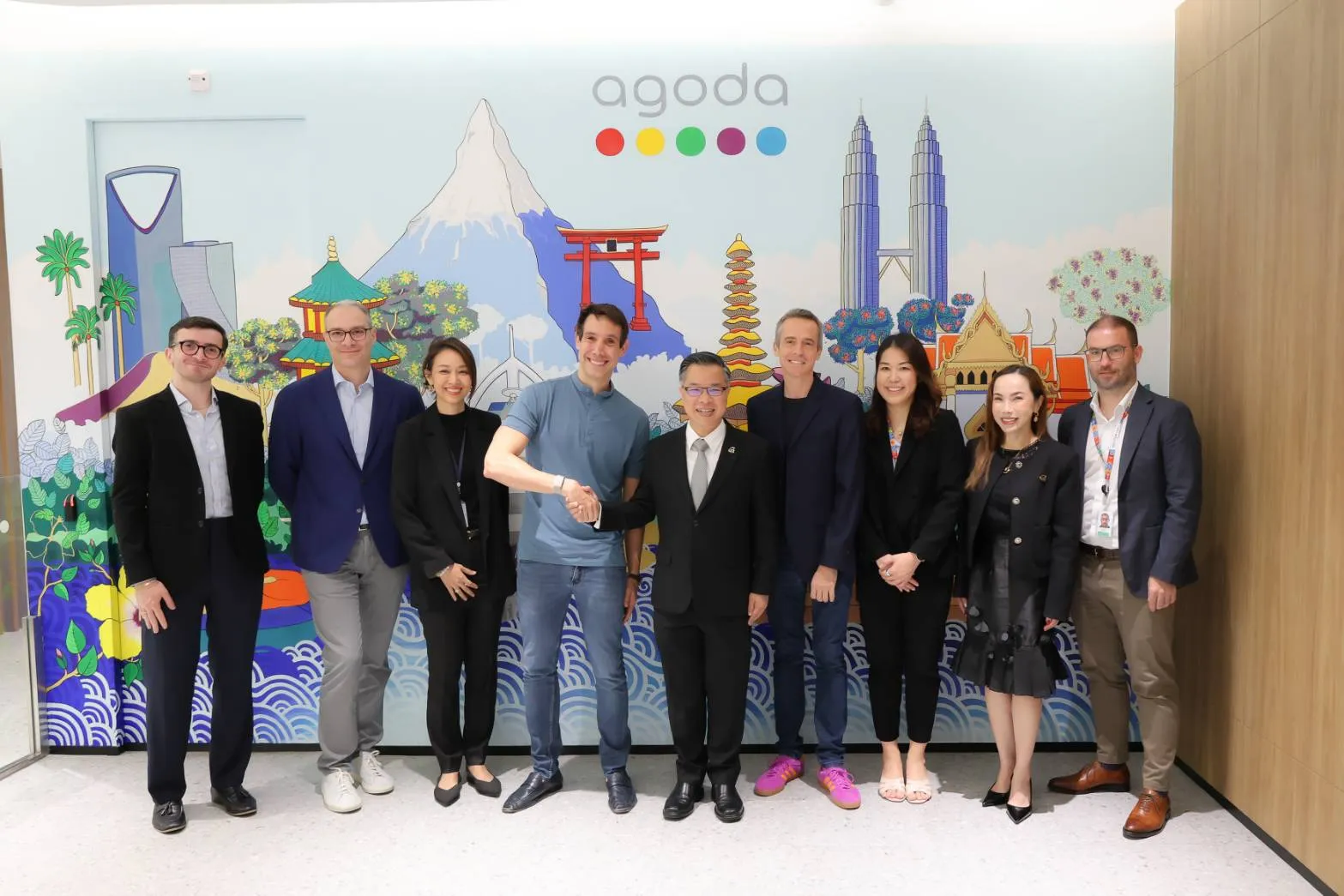 ผู้บริหารชายหญิง 9 คนยืนถ่ายภาพร่วมกันหน้าผนังโลโก้ Agoda ที่ตกแต่งด้วยภาพวาดสถานที่สำคัญระดับโลก โดยมีสองคนตรงกลางจับมือทักทายกัน แสดงถึงความร่วมมือและการลงทุนขยายฐานการจ้างงานด้านเทคโนโลยีของ Agoda ในประเทศไทย
