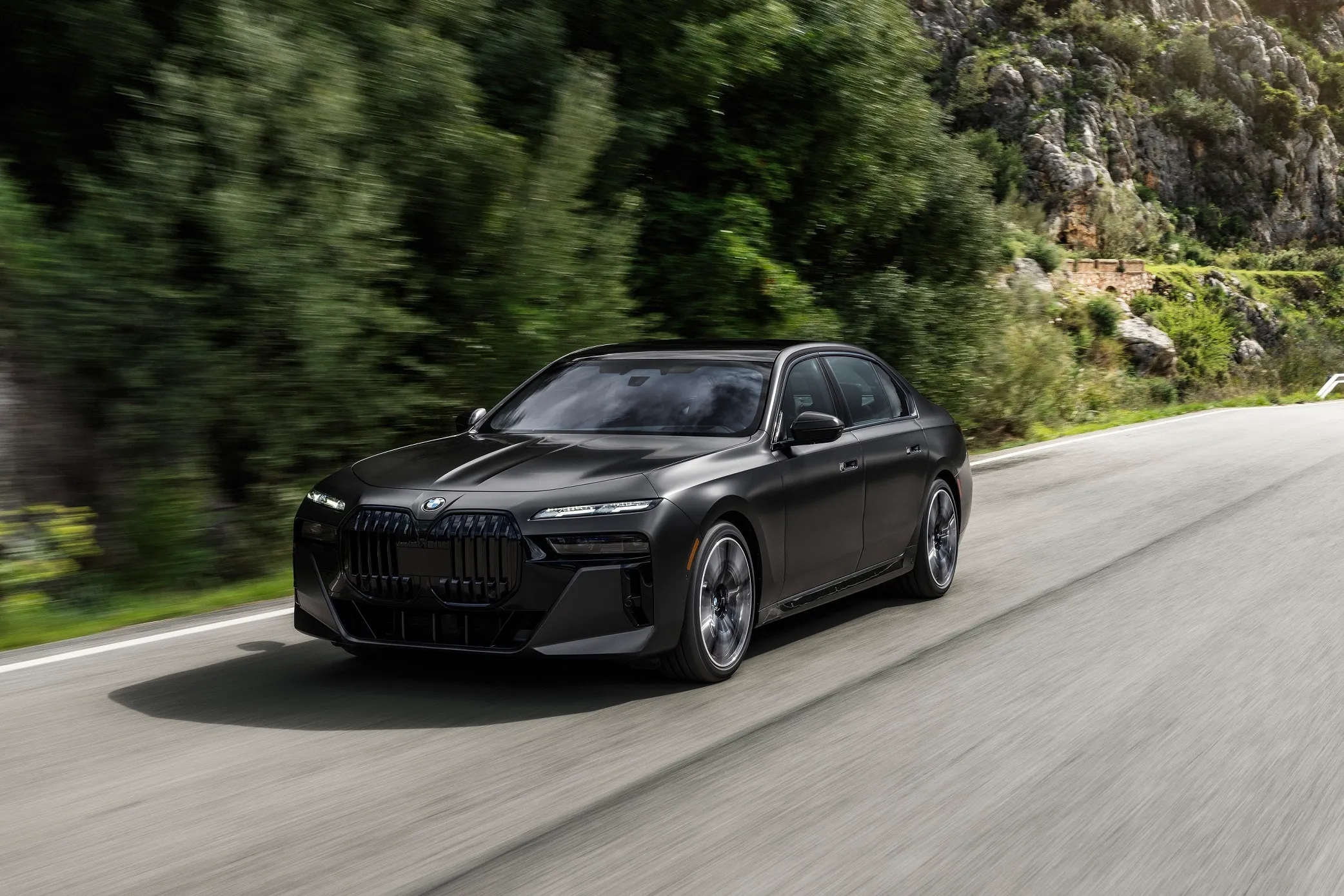 รถยนต์ BMW ซีดานสีดำกำลังแล่นด้วยความเร็วบนถนนลาดยางท่ามกลางทิวทัศน์ภูเขาและป่าไม้เขียวขจี