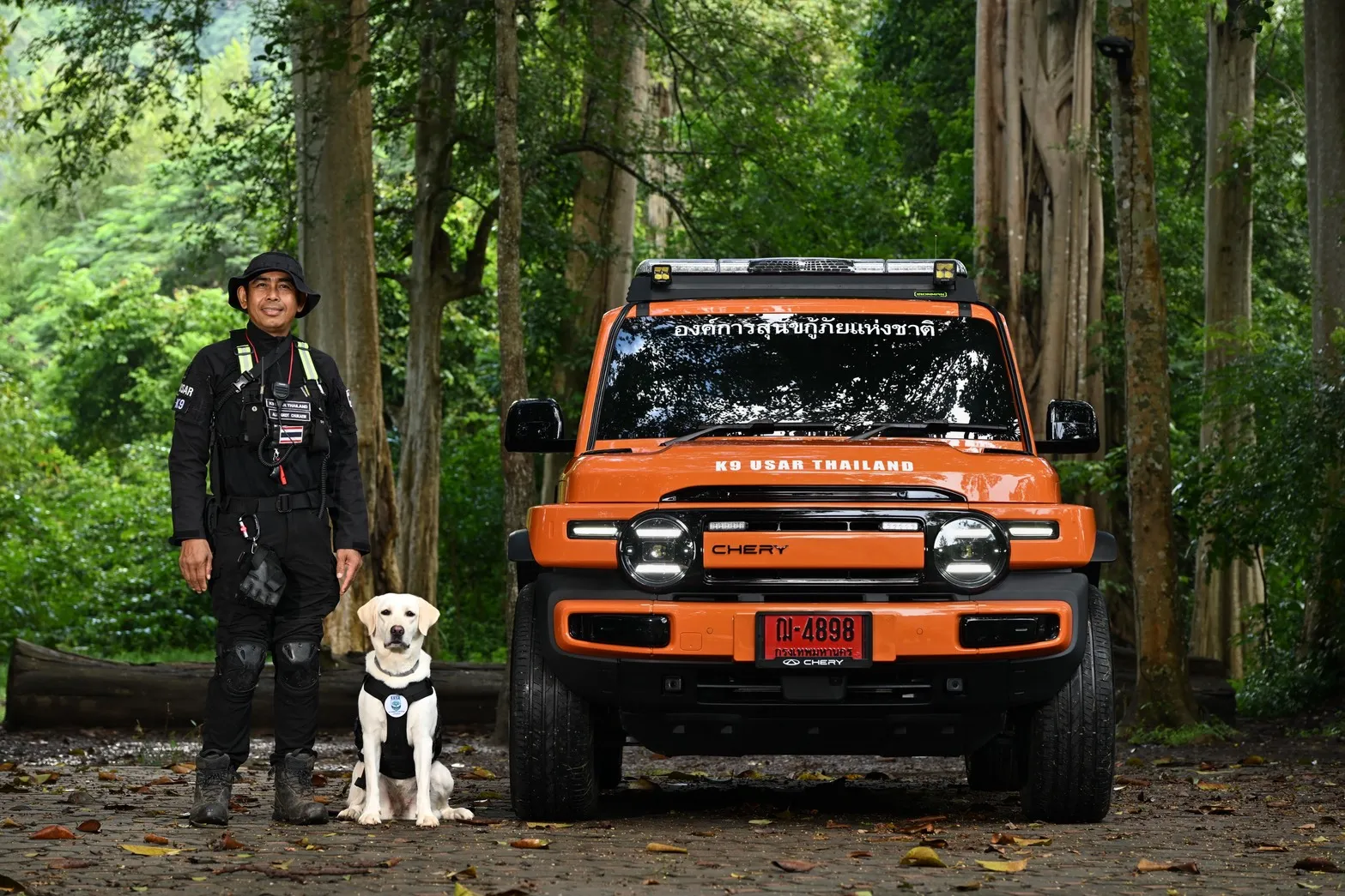 เจ้าหน้าที่หน่วยกู้ภัยพร้อมสุนัข K9 ยืนอยู่หน้ารถยนต์ไฟฟ้า Chery V23 สีส้มรุ่น K9 USAR Thailand ท่ามกลางป่าไม้เขียวขจี