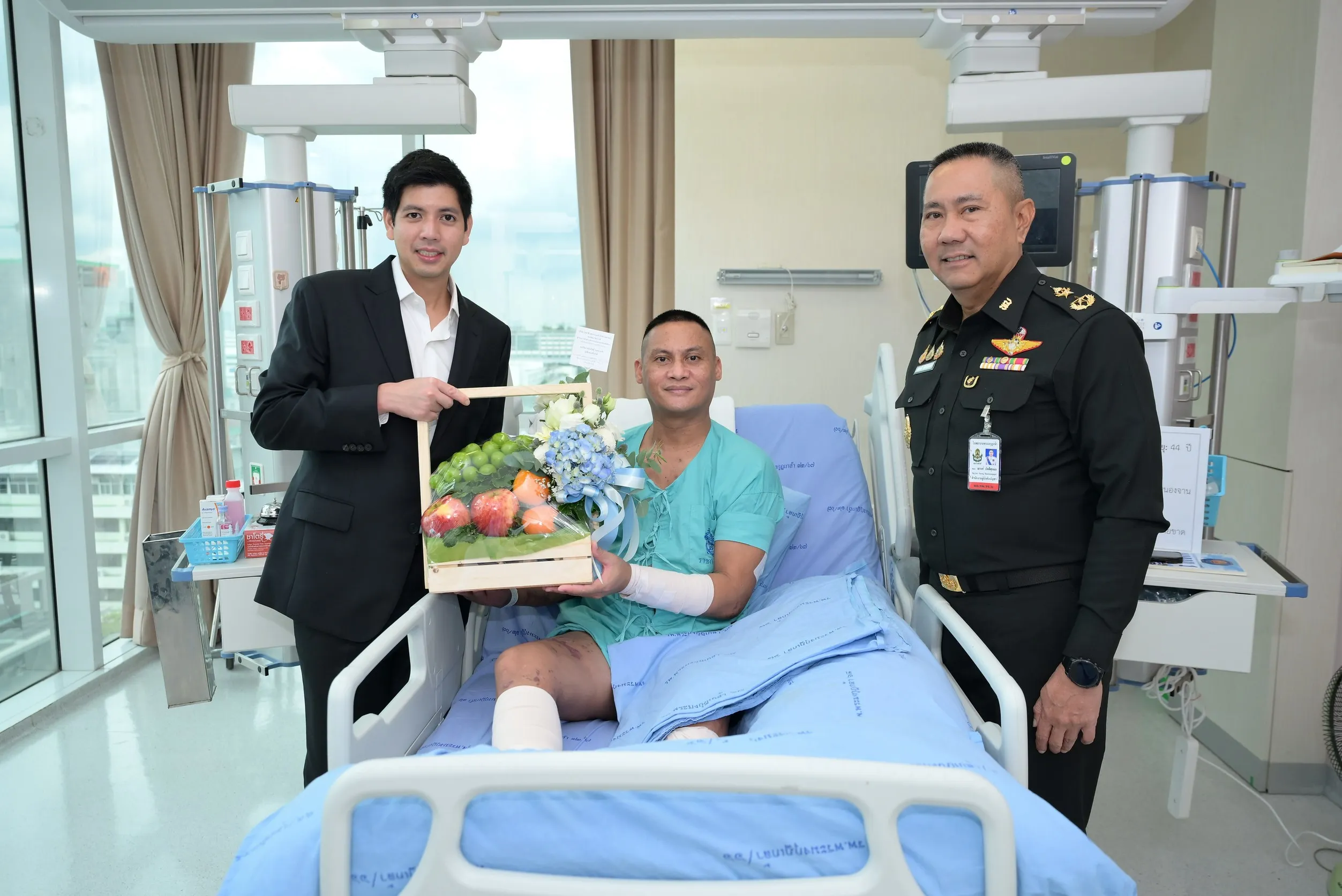 ผู้บริหารในชุดสูทและนายทหารยศสูงยืนมอบกระเช้าผลไม้และดอกไม้ให้ทหารบาดเจ็บที่นั่งอยู่บนเตียงผู้ป่วยในโรงพยาบาล เพื่อเป็นกำลังใจจากภารกิจปกป้องชายแดนไทย-กัมพูชา