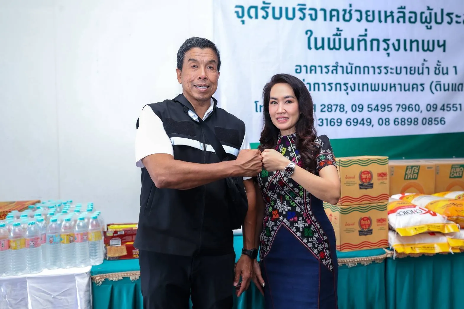 ตัวแทนเอส แอนด์ พี และเจ้าหน้าที่กรุงเทพมหานครชนหมัดกันต่อหน้าจุดรับบริจาคสิ่งของช่วยเหลือผู้ประสบภัยน้ำท่วม พร้อมกล่องผลิตภัณฑ์และน้ำดื่มที่จัดเตรียมไว้บริจาค