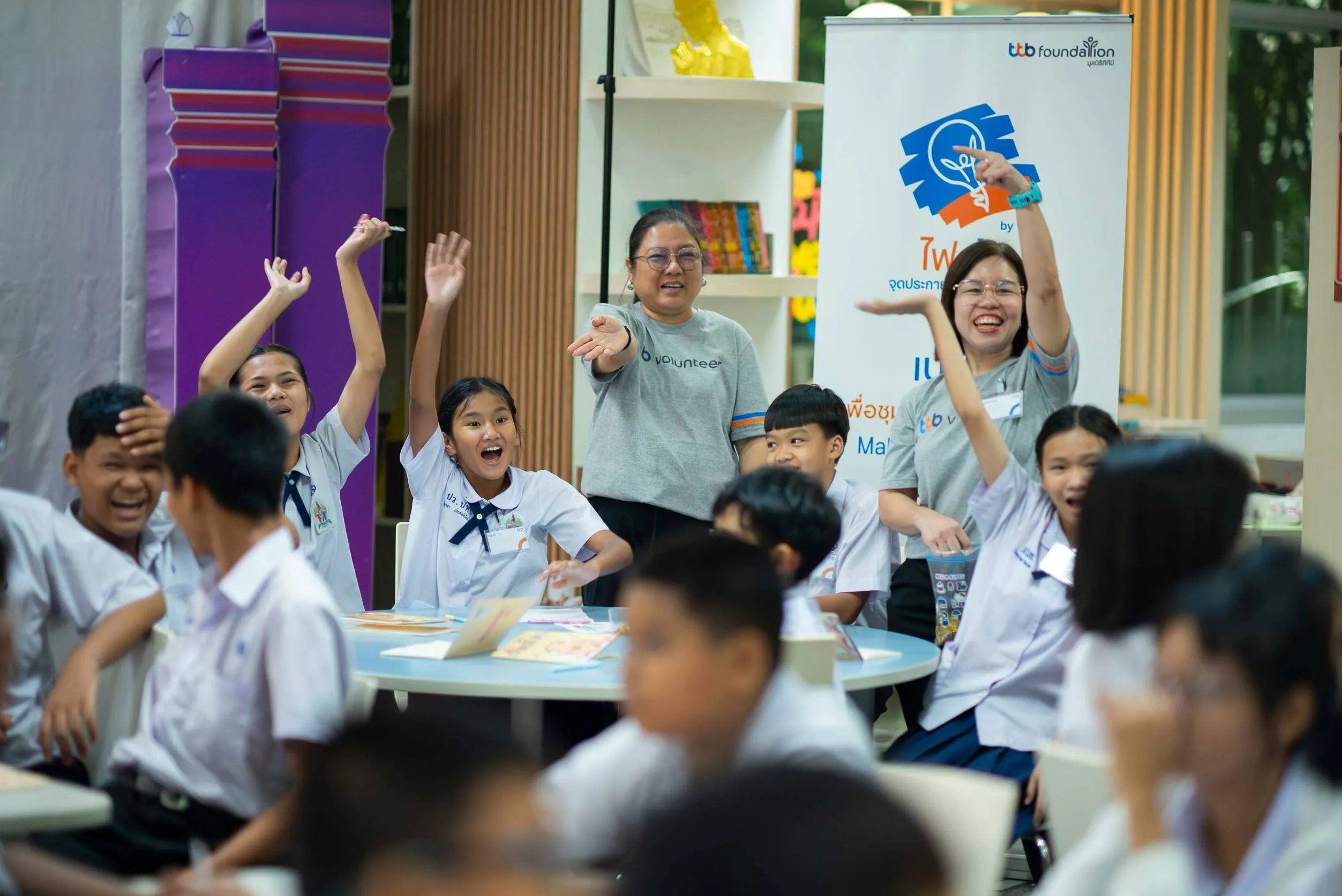 อาสาสมัครจากทีทีบีทำกิจกรรมสนุกสนานร่วมกับนักเรียนในห้องเรียน โดยทุกคนยกมือและยิ้มแย้มอย่างมีความสุขภายใต้บรรยากาศโครงการ "รุ่นพี่การเงิน" ของมูลนิธิทีทีบี