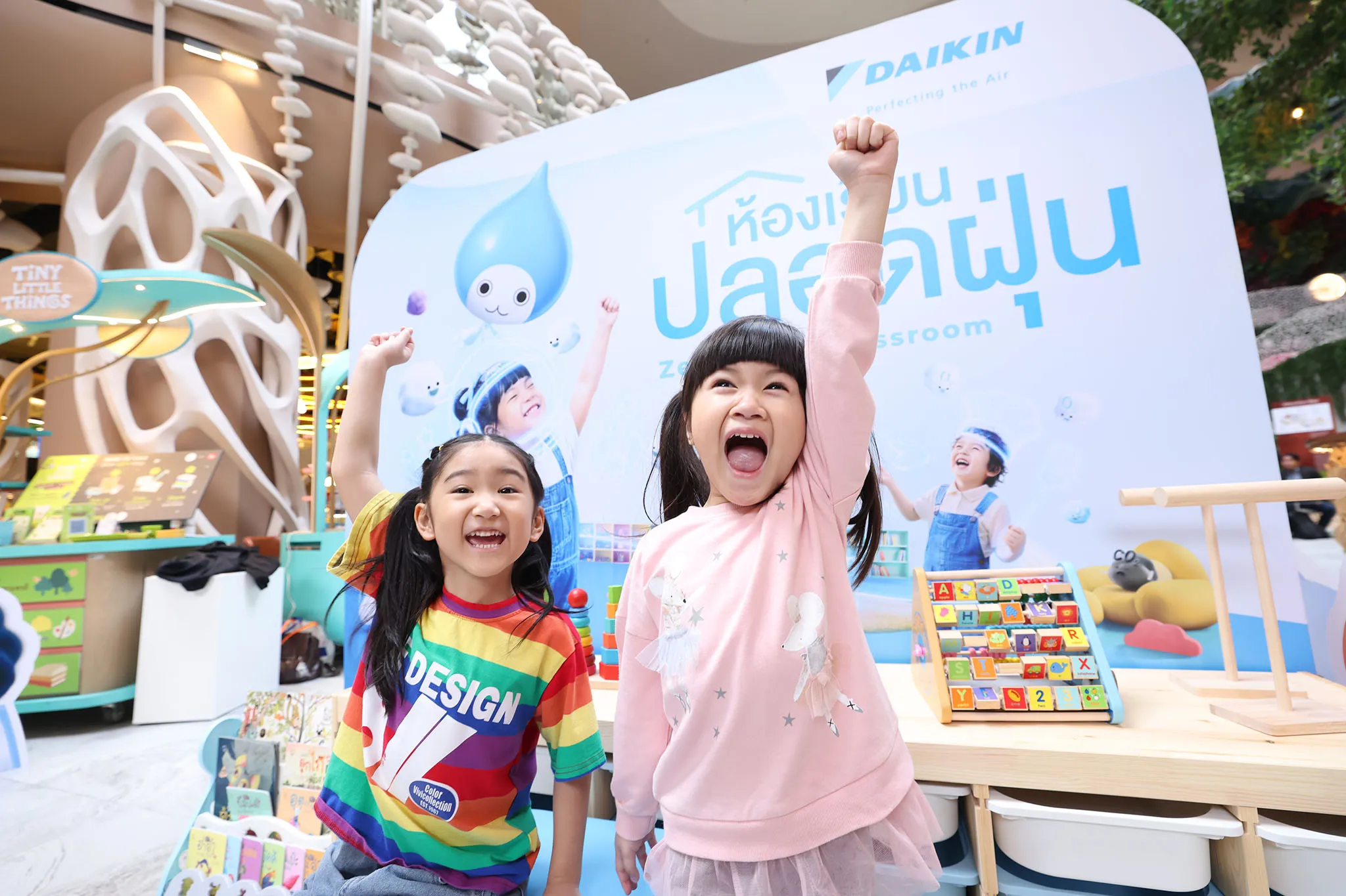 เด็กหญิงสองคนยิ้มร่าเริงยกมือเชียร์หน้าป้ายประชาสัมพันธ์โครงการ "ห้องเรียนปลอดฝุ่น" ของไดกิ้น ภายในพื้นที่กิจกรรมเด็กที่ตกแต่งด้วยของเล่นและหนังสือสีสันสดใส