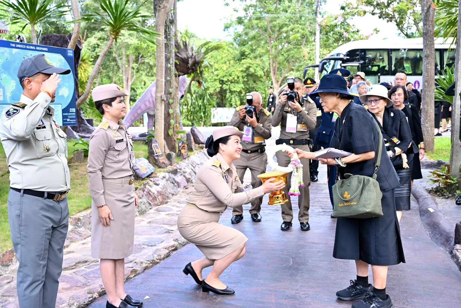 เจ้าหน้าที่ตำรวจหญิงในเครื่องแบบคุกเข่าน้อมถวายพานดอกไม้แด่สตรีสูงอายุ ท่ามกลางคณะเจ้าหน้าที่และนักท่องเที่ยวที่ยืนรอต้อนรับบริเวณทางเดินอุทยานไดโนเสาร์ภูเวียง