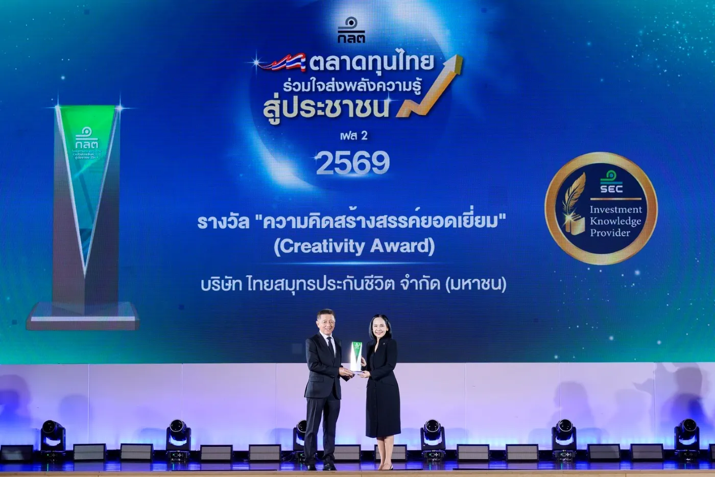 ผู้บริหารชายและหญิงถ่ายภาพร่วมกันบนเวทีขณะรับมอบรางวัล Creativity Award จากโครงการตลาดทุนไทยร่วมใจส่งพลังความรู้สู่ประชาชน เฟส 2 ประจำปี 2569 ซึ่งมอบให้แก่บริษัท ไทยสมุทรประกันชีวิต จำกัด (มหาชน) โดยมีถ้วยรางวัลสีเขียวและโลโก้ ก.ล.ต. Investment