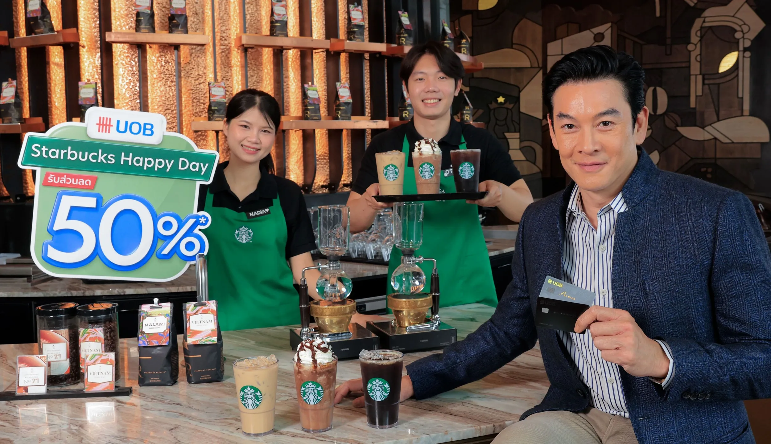 ผู้บริหารชายถือบัตร UOB ยืนอยู่หน้าเคาน์เตอร์สตาร์บัคส์พร้อมพนักงานบาริสต้าสองคนและป้ายโปรโมชั่น UOB Starbucks Happy Day ลดส่วนลด 50% ท่ามกลางเครื่องดื่มกาแฟและเมล็ดกาแฟที่จัดวางบนเคาน์เตอร์