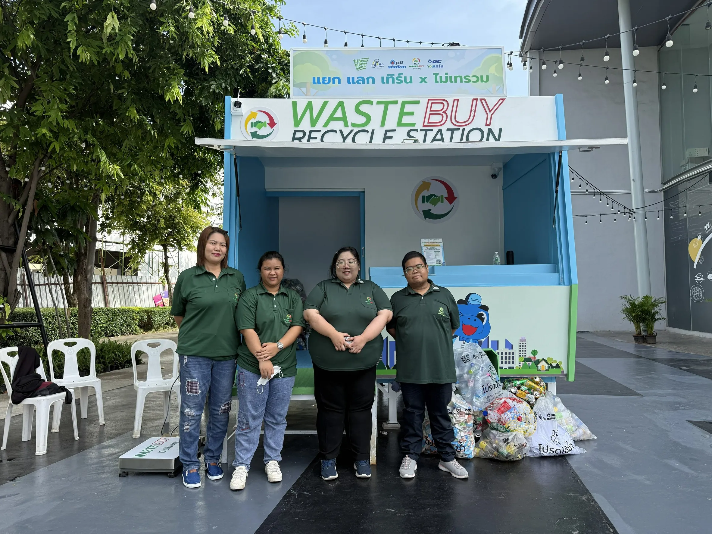 เจ้าหน้าที่ 4 คนสวมเสื้อโปโลสีเขียวยืนถ่ายรูปหน้าตู้ WasteBuy Recycle Station พร้อมถุงขยะรีไซเคิลประเภทขวดพลาสติกและกระป๋องวางอยู่ด้านข้าง